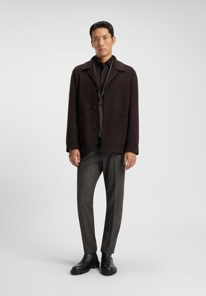 Abrigo de mezcla de lana marrón con grandes botones y dos bolsillos frontales, combinado con una camisa oscura y pantalones grises texturizados. Botas negras hasta el tobillo completan el look.