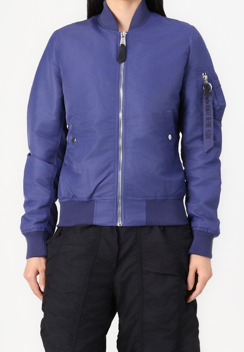 Personne portant une veste bomber bleue avec une fermeture éclair à l'avant, des poignets côtelés et un pantalon noir, debout sur un fond blanc.