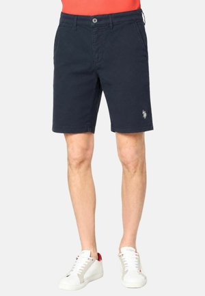 Uomo che indossa pantaloncini blu navy con tasche anteriori e scarpe da ginnastica bianche, in piedi su uno sfondo neutro.
