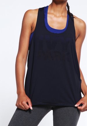 Vrouw draagt een navy tanktop met de tekst "IVY PARK" over een blauwe sportbeha en grijze legging, houdt de zoom van de tanktop vast.