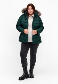 Giacca imbottita verde con cappuccio foderato in pelliccia, caratterizzata da un motivo a righe orizzontali, zip frontale e due tasche laterali. Indossata sopra un maglione leggero.