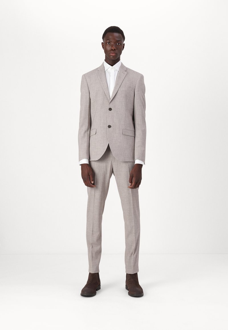 Fato cinza claro, ajustado, com lapela notched, dois botões e bolsos laterais. Camisa branca por baixo, complementada por sapatos castanho-escuros.