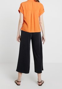 Pomarańczowa koszula z krótkim rękawem i plisą z tyłu, zestawiona z czarnymi szerokimi spodniami. Cały look cechuje się luźnym krojem i miękką fakturą.