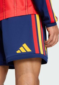 Sportovní kraťasy Adidas se základní modrou barvou a červenými a žlutými svislými pruhy. Na boku je logo Adidas a emblém Španělska.