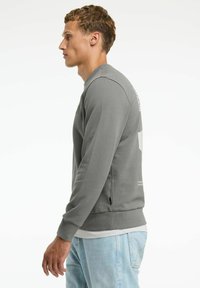 Felpa grigia a maniche lunghe con texture a coste, caratterizzata da spacco laterale, colletto a girocollo e dettagli con logo sulla schiena. Indossata sopra jeans blu chiaro.
