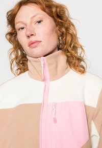 Casaco de fleece com gola alta, design em blocos de cor em bege, rosa e creme, com dois fechos frontais e uma textura suave.