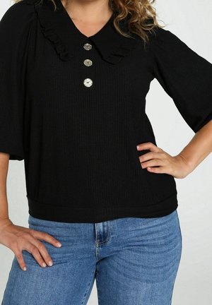 Femme portant une blouse noire texturée avec col à volants et trois boutons métalliques, associée à un jean bleu, mains sur les hanches.