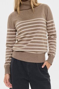 Kobieta ubrana w brązowy sweter z golfem z beżowymi poziomymi paskami, zestawiony z luźnymi czarnymi spodniami, jedna ręka w kieszeni.