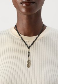 Necklace od crnih i sivih perlica s zlatnim privjeskom u obliku perja, nosi se preko rebraste bež majice. Tekstura i boje stvaraju upečatljiv kontrast.