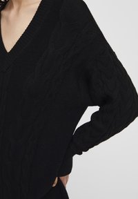 Maglione nero lavorato a maglia con scollo a V e motivo a trecce testurizzato sulle maniche. Presenta dettagli a costine sui polsini e sull'orlo.
