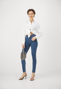 Vit knuten skjorta, blå slim-fit jeans, svarta klackar med remmar och en liten mönstrad handväska. Enkelt och rent utseende.