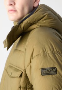 Oliwkowa kurtka puchowa z zamkiem błyskawicznym z przodu, kapturem i kieszonką na piersi; posiada czarną naszywkę z logo "Barbour International" na rękawie.
