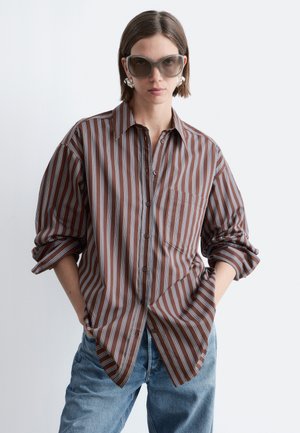 Donna con camicia marrone a righe oversize, jeans blu, grandi occhiali da sole quadrati e orecchini vistosi, in piedi con le mani nelle tasche.