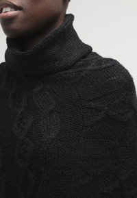 Personne portant un pull à col roulé noir en maille torsadée, mettant en valeur les motifs tricotés et les détails texturés du tissu sur l'épaule et le cou.