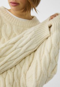 Roomkleurige kabelgebreide trui met een brede halslijn en geribbelde manchetten. Getextureerde details voegen diepte toe, gemaakt van zachte, dikke garen.
