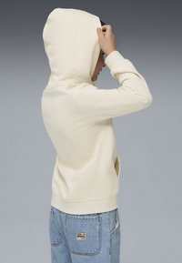 Felpa con cappuccio color crema realizzata in tessuto morbido, con un design aderente e polsini a coste. Indossata con jeans denim chiari.