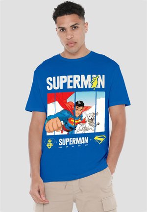 Young man wearing a blue Superman t-shirt featuring Superman flying with a dog and text "Look up in the sky" on a plain background.