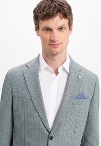 Hellgrauer Blazer mit feinem strukturierten Muster, Reverskragen, einem Knopf und einem blauen Taschentuch. Getragen über einem weißen Hemd.