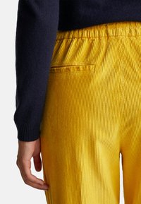 Gele corduroy broek met een ribbeltextuur, voorzien van een elastische tailleband en een klein achterzakje. De hand van het model rust op de broek.