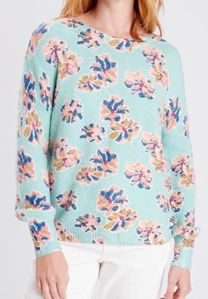Kvinde iført lyseblå sweater med lyserøde, blå og beige blomsterprint, kombineret med hvide bukser, vist fra nakke til hofter.