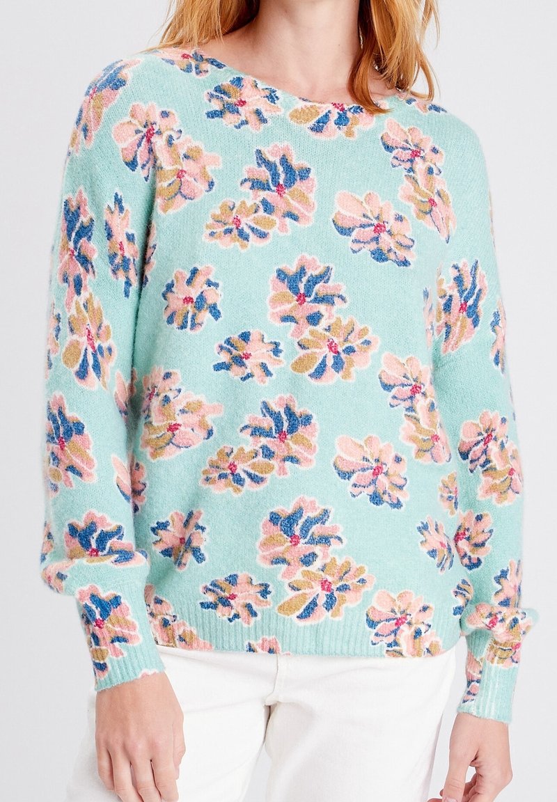 Femme portant un pull bleu clair avec des motifs floraux roses, bleus et beiges, associé à un pantalon blanc, vue du cou jusqu’aux hanches.