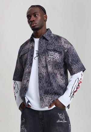 Jeune homme aux cheveux tressés portant une chemise noire et grise à manches courtes à motifs par-dessus une chemise blanche à manches longues avec des manches graphiques et un jean noir brodé.
