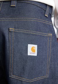 Tumši denima džinsi ar dzeltenām šuvēm, redzamu aizmugures kabatu un baltu etiķeti ar dzeltenu logotipu un sarkanu tekstu.