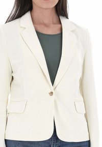 Blazer bianco con vestibilità sartoriale, dotato di chiusura con un solo bottone, due tasche e un tessuto texture. Indossato sopra una maglietta verde.