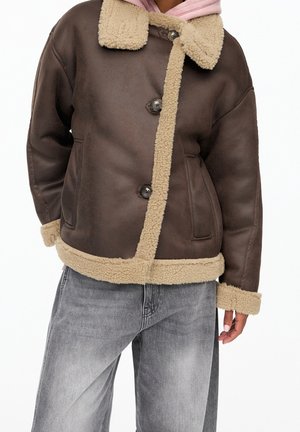 Personne portant une veste en peau lainée marron foncé avec une bordure en polaire beige et un jean gris, debout devant un fond blanc uni.