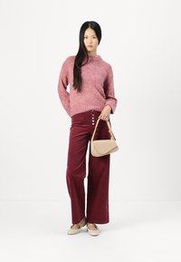 Pantaloni a gamba larga in velluto rosso, maglione rosa lavorato a maglia, borsa beige e scarpe chiare. Il maglione ha una consistenza spessa e un colletto arrotolato.