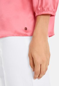 Rosa Satinbluse mit Puffärmeln. Verfügt über ein kleines, rundes Logodetail am Saum. Kombiniert mit weißen Stoffhosen.
