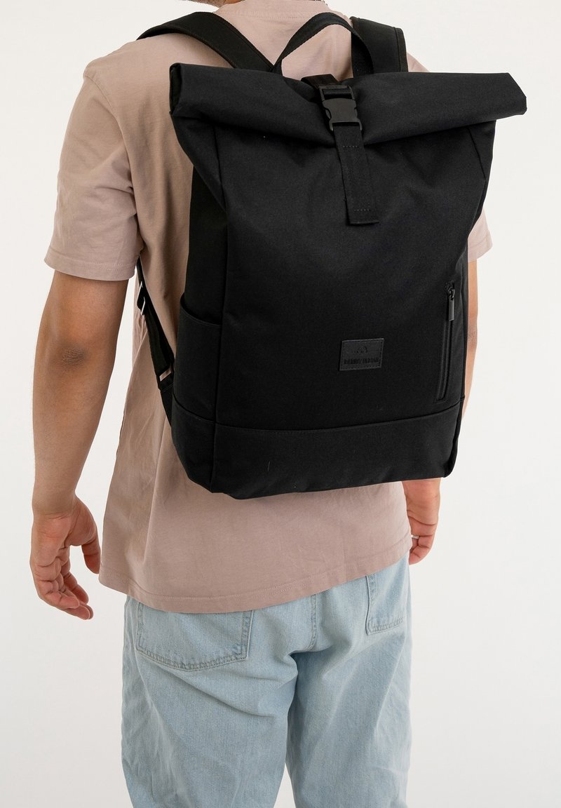 Person wearing a black roll-top backpack with side pockets, zippered front pocket, and adjustable shoulder straps over a beige shirt and light jeans.