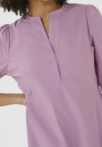 Jasnofioletowa tunika z kołnierzykiem mandaryńskim, pojedynczą listwą na guziki i krótkimi bufkowymi rękawami. Gładka tekstura i prosty design.