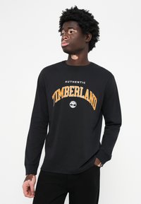 Crna majica s dugim rukavima s okruglim izrezom, hrabar narančasti natpis "TIMBERLAND" i mali logotip na središtu. Materijal: pamuk.