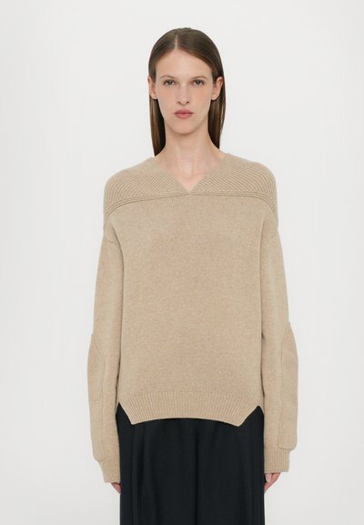 Beige strikket sweater med V-udskæring, ribbestrikkede detaljer ved skuldrene og brede ærmer, der har en afslappet pasform og delte sømme.