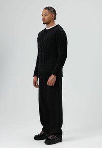 Zwarte gebreide trui met een gestructureerd ruitpatroon, gecombineerd met losse zwarte broek en robuuste zwarte schoenen met roze veters.