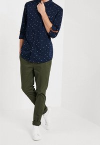 Marineblå skjorte med knapper og hvide og røde polkaprikker; ærmerne er rullede op; kombineret med grønne bukser og hvide sneakers.