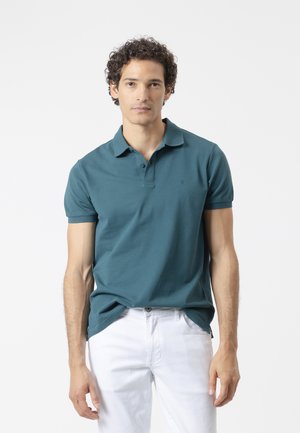 Hombre con pelo rizado que lleva una camiseta polo de manga corta en color verde azulado y pantalones blancos, de pie frente a un fondo claro y liso.