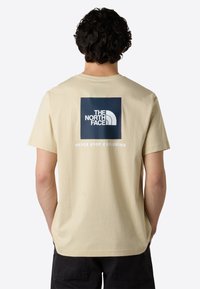 Béžové bavlněné tričko s velkým námořnickým čtvercovým logem a bílým textem na zádech, které zní "THE NORTH FACE" a "NEVER STOP EXPLORING."