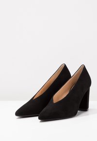 Coppia di scarpe nere in suede con tacco alto, punte appuntite e tacco a blocco su una superficie bianca contro uno sfondo semplice.