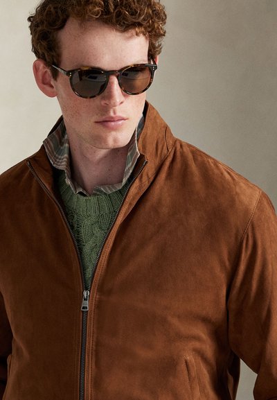 Tânăr cu păr roșcat și creț, purtând ochelari de soare cu rame în model tartarugă, jachetă maro din piele întoarsă, pulover verde tricotat și gulerul cămășii în carouri vizibil.