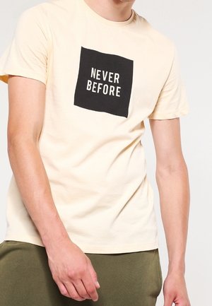 Osoba nosząca beżowy T-shirt z czarnym kwadratem na klatce piersiowej, zawierający biały napis "NEVER BEFORE", w połączeniu z oliwkowozielonymi spodniami.