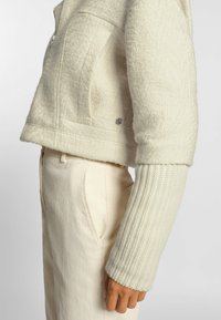 Sido vy av en person som bär en off-white texturerad jacka med stickade ärmar och ljusa kordbyxor, handen avslappnad vid sin sida.