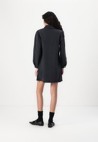 Rochie din denim negru cu guler și mâneci lungi evazate, având un croi lejer și un tiv destrămat. Poartă șosete negre și pantofi cu model.