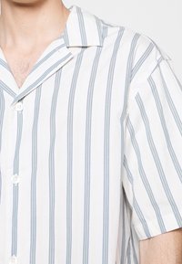 Homme portant une chemise blanche à manches courtes avec des rayures verticales bleues et grises et des boutons blancs, col ouvert au niveau du cou.