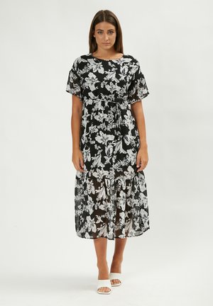 Vestido negro con estampado floral en blanco, mangas cortas, cintura ceñida, dobladillo en capas, hecho de una tela ligera y transparente. Un par de zapatos blancos.