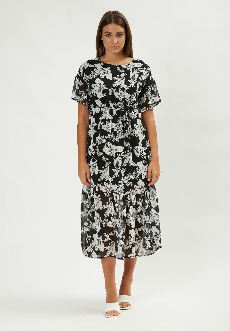 Schwarzes Blumenkleid mit weißen Blumenmustern, kurzen Ärmeln, tailliertem Schnitt, geschichteter Saum, aus leichtem, transparentem Stoff. Ein Paar weiße Schuhe.