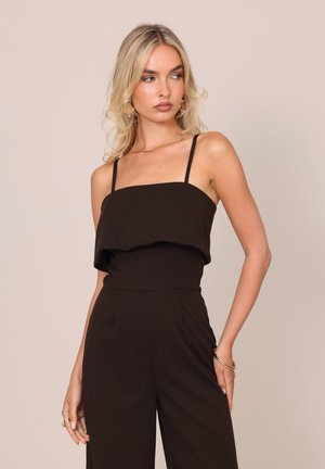 Femme blonde portant une combinaison noire sans manches avec des bretelles fines et des bijoux dorés sur un fond beige uni.