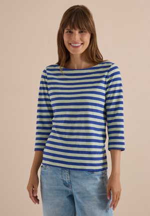 Femme aux cheveux bruns portant une chemise à manches longues rayée bleu et vert clair et un jean bleu clair, souriant à la caméra.