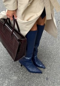 Stivali blu in pelle con texture coccodrillo, alti fino al ginocchio, con punte appuntite e tacchi a stiletto, abbinati a una borsa a tracolla in pelle marrone scuro.